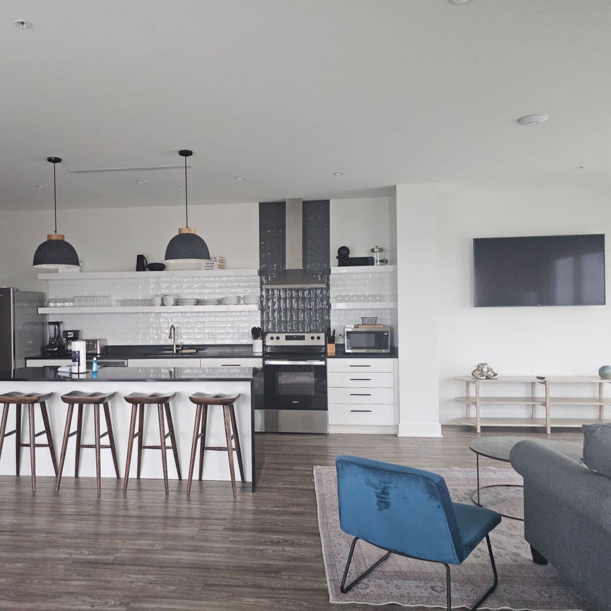 Open kitchen area with stools at a counter, living room area with blue chair and grey couch, flatscreen tv on the wall above a low shelving unit