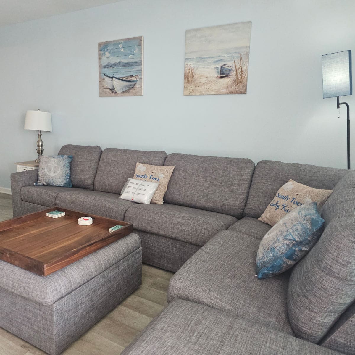 grey couch with a few pillows on the couch, beach art on the wall, ottoman in the middle