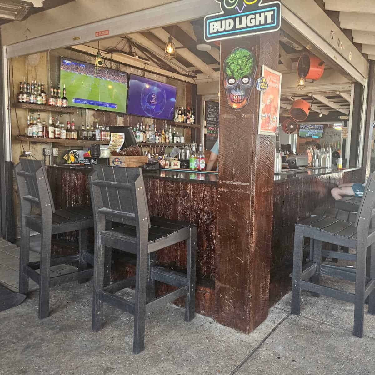 outdoor covered bar with tall cairs, bottles lined on shelves near two tv's