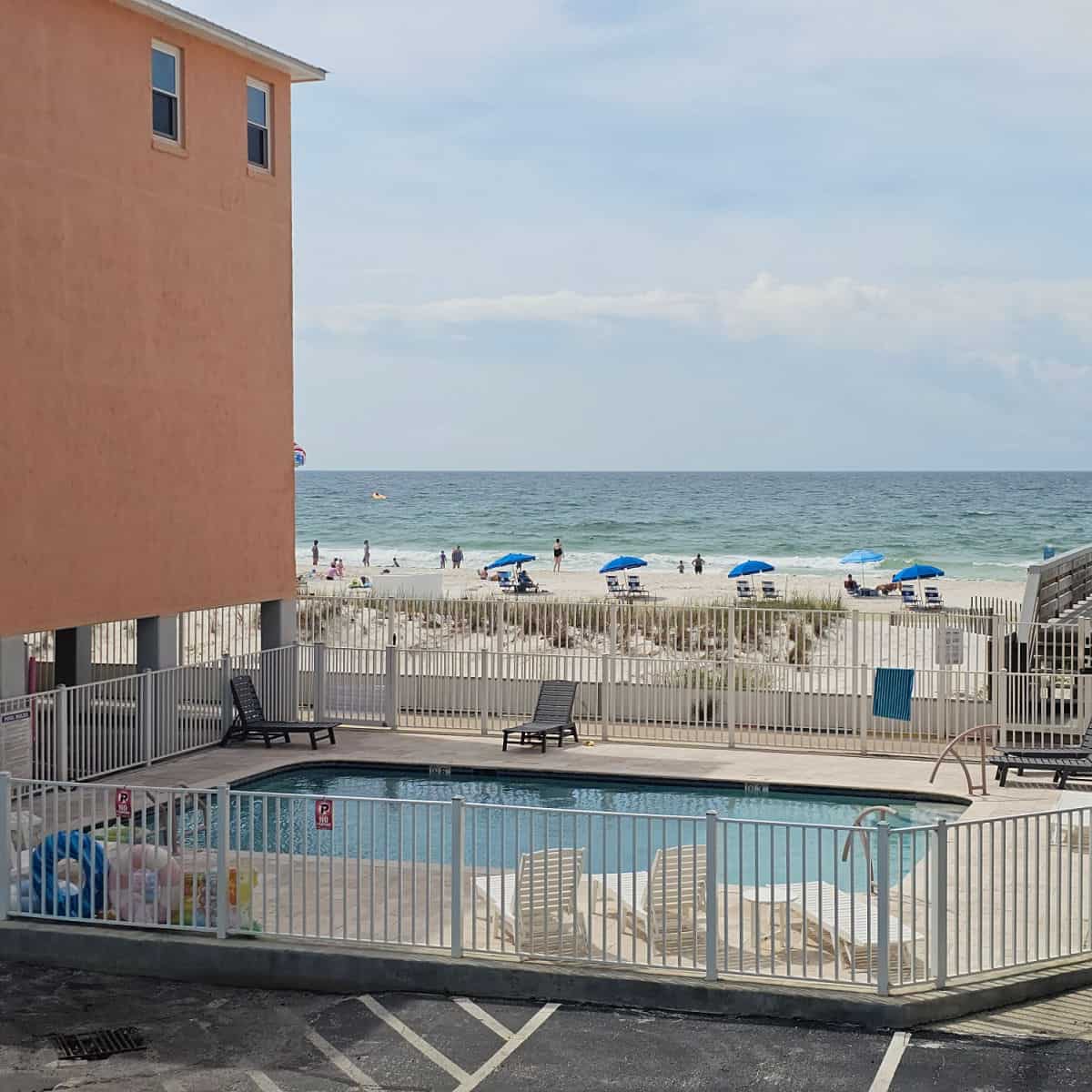 outdoor pool with beach chairs behind a white fence next to a 2 story building, gulf and beach in the background.
