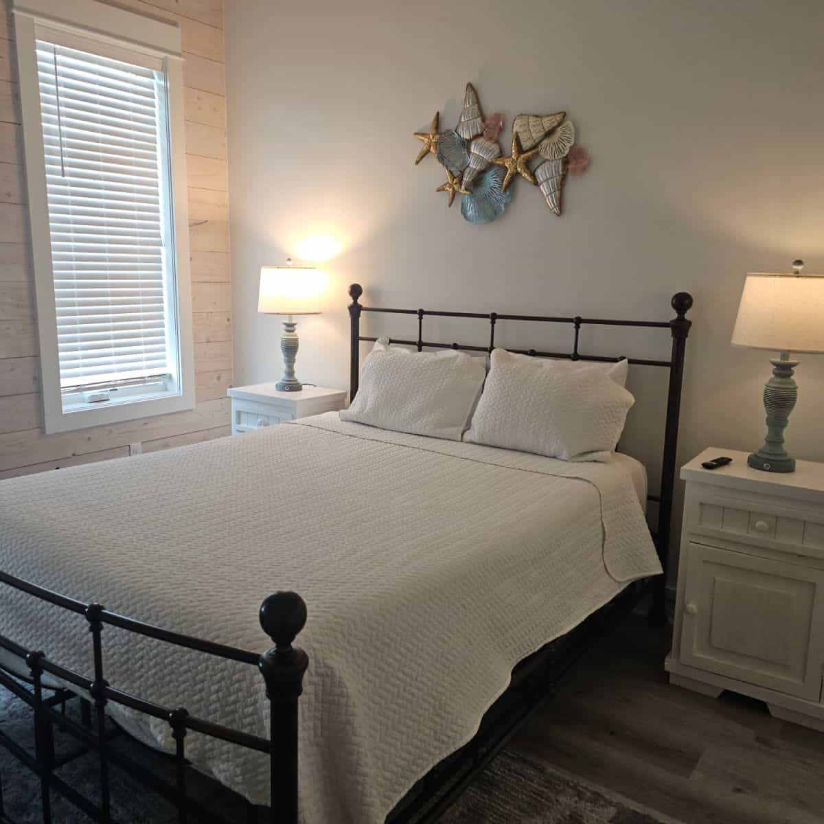bedroom with metal frame bed, white linens, white nightstands with lamps on them, shell art on the wall