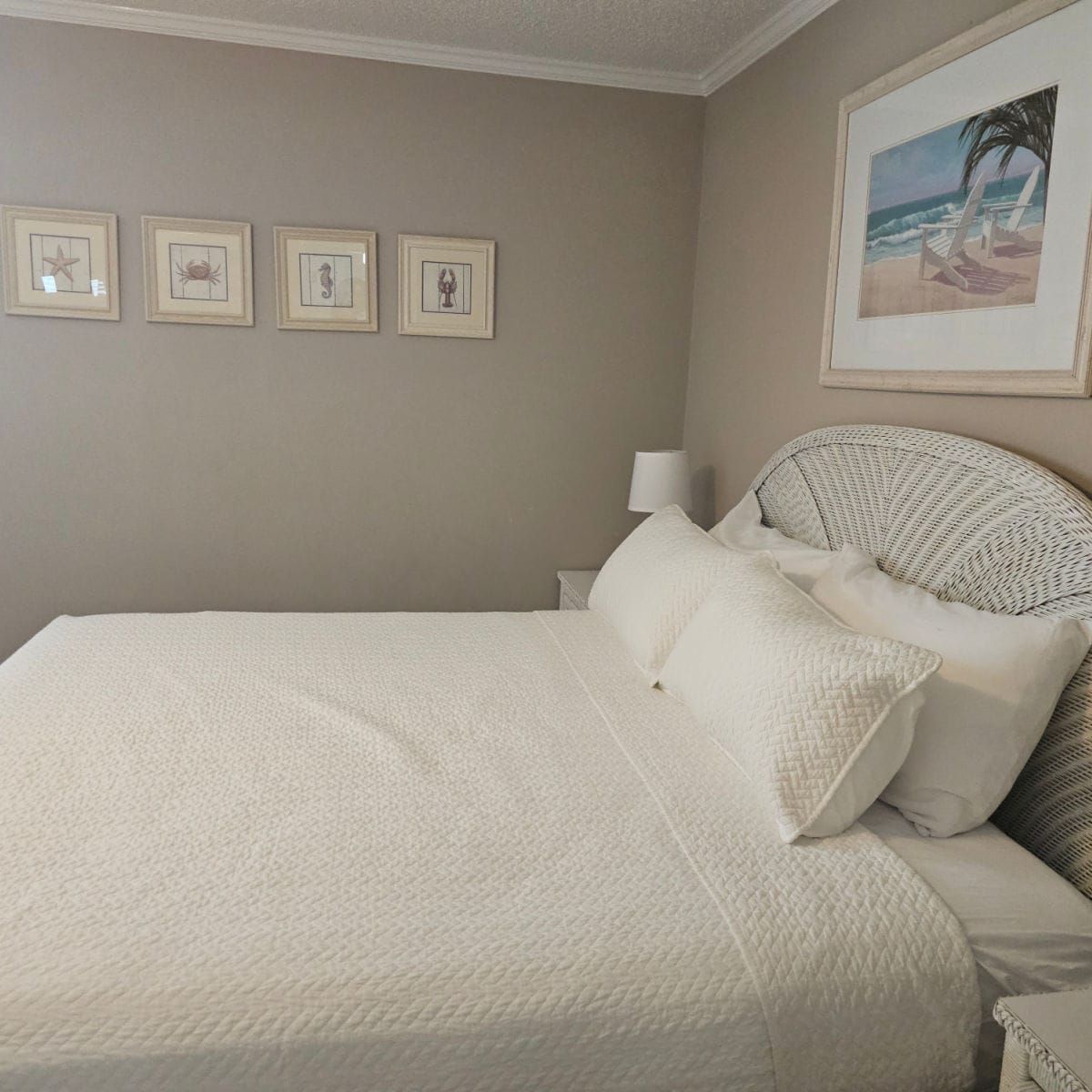 bed with rattan headboard, two nightstands one with a white lamp, tropical art print over the bed, and four coastal designed art prints on the other wall