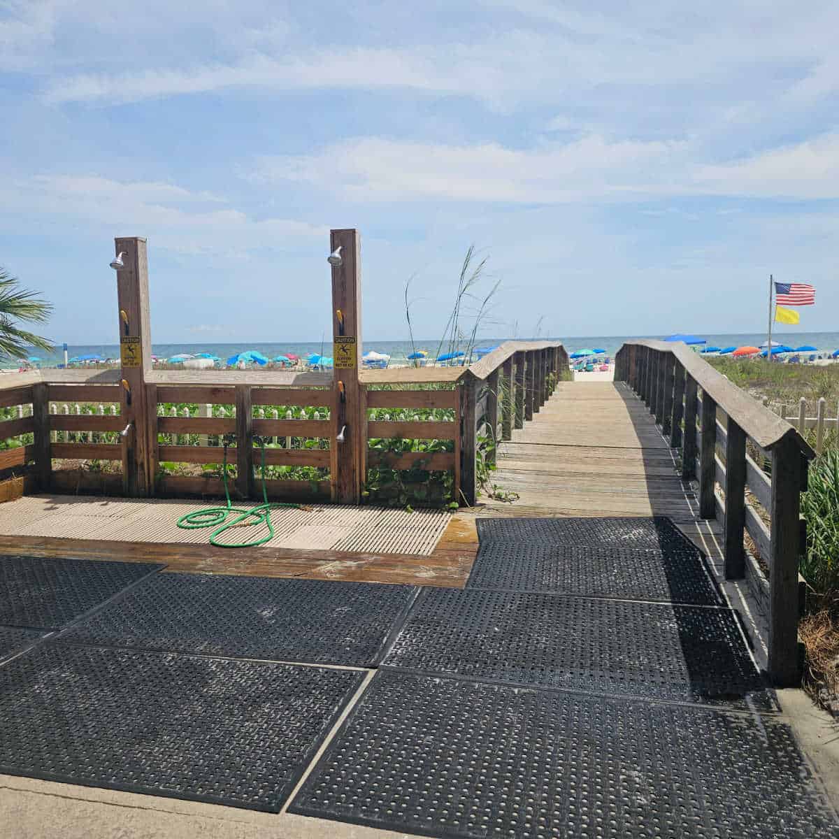 wooden boardwalk over sand dunes, two outdoor showers, american flag and yellow flag blowing in the wind, umbrellas along the beach line