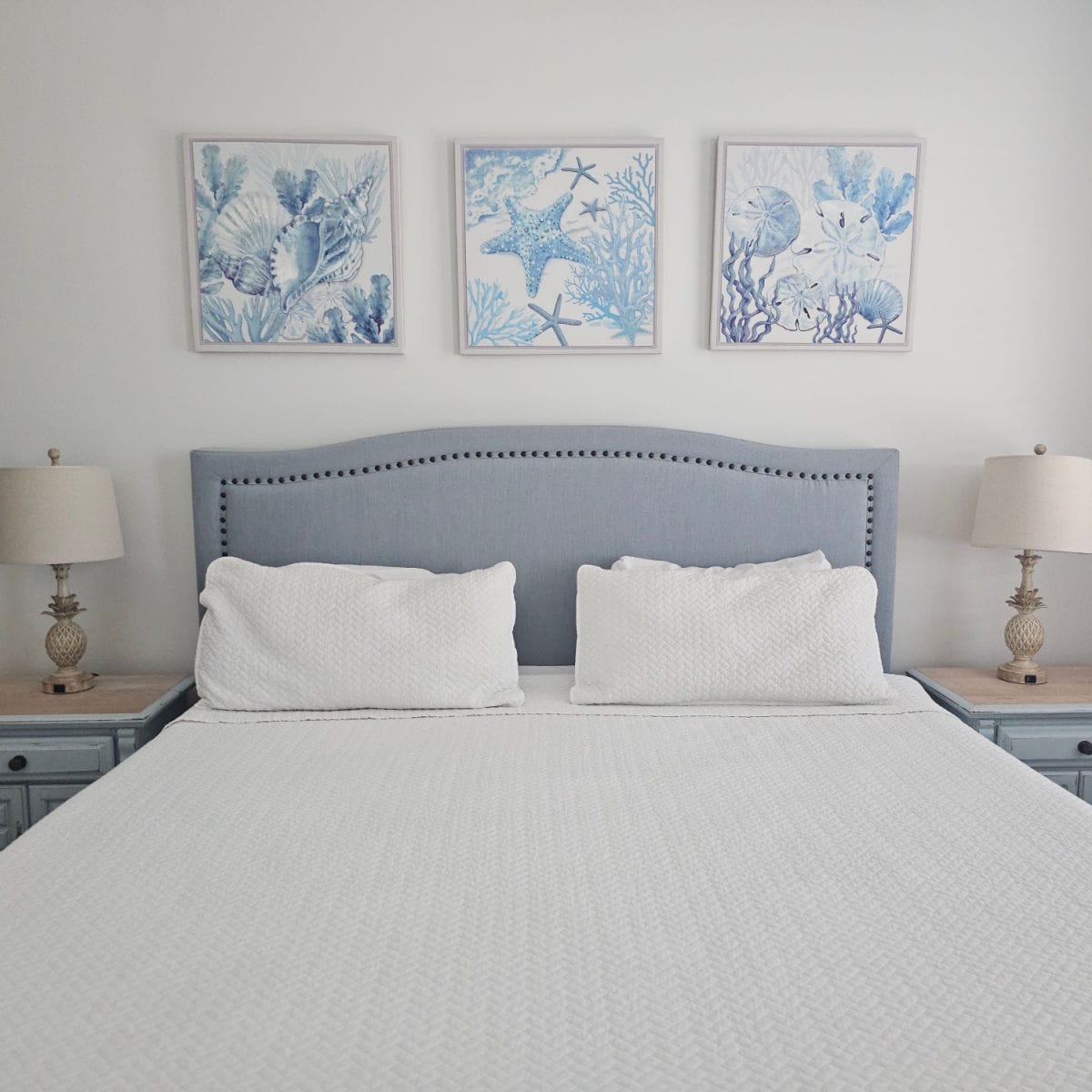 Bedroom with king bed, white linens, seashell art on the wall, nightstands with pineapple shaped base on lamps
