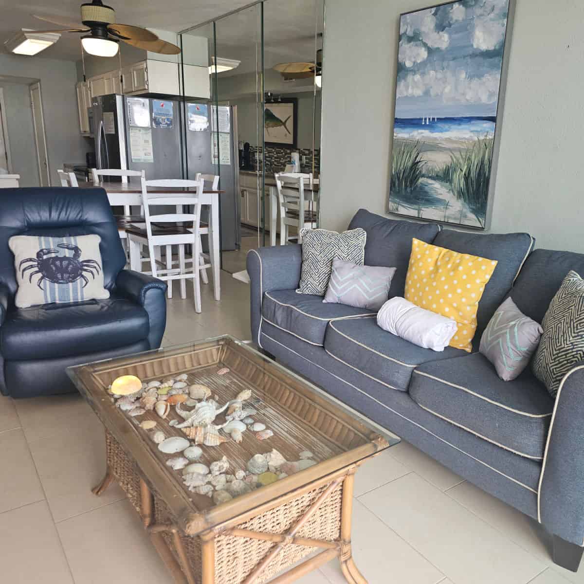 Living room with blue grey couch with lots of pillows, shells in a glass topped table, blue leather chair with crab pillow