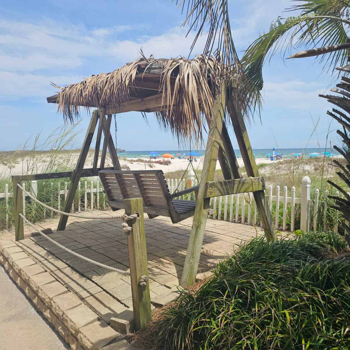 wooden swing with thatch covering looking out to the beach with umbrellas.