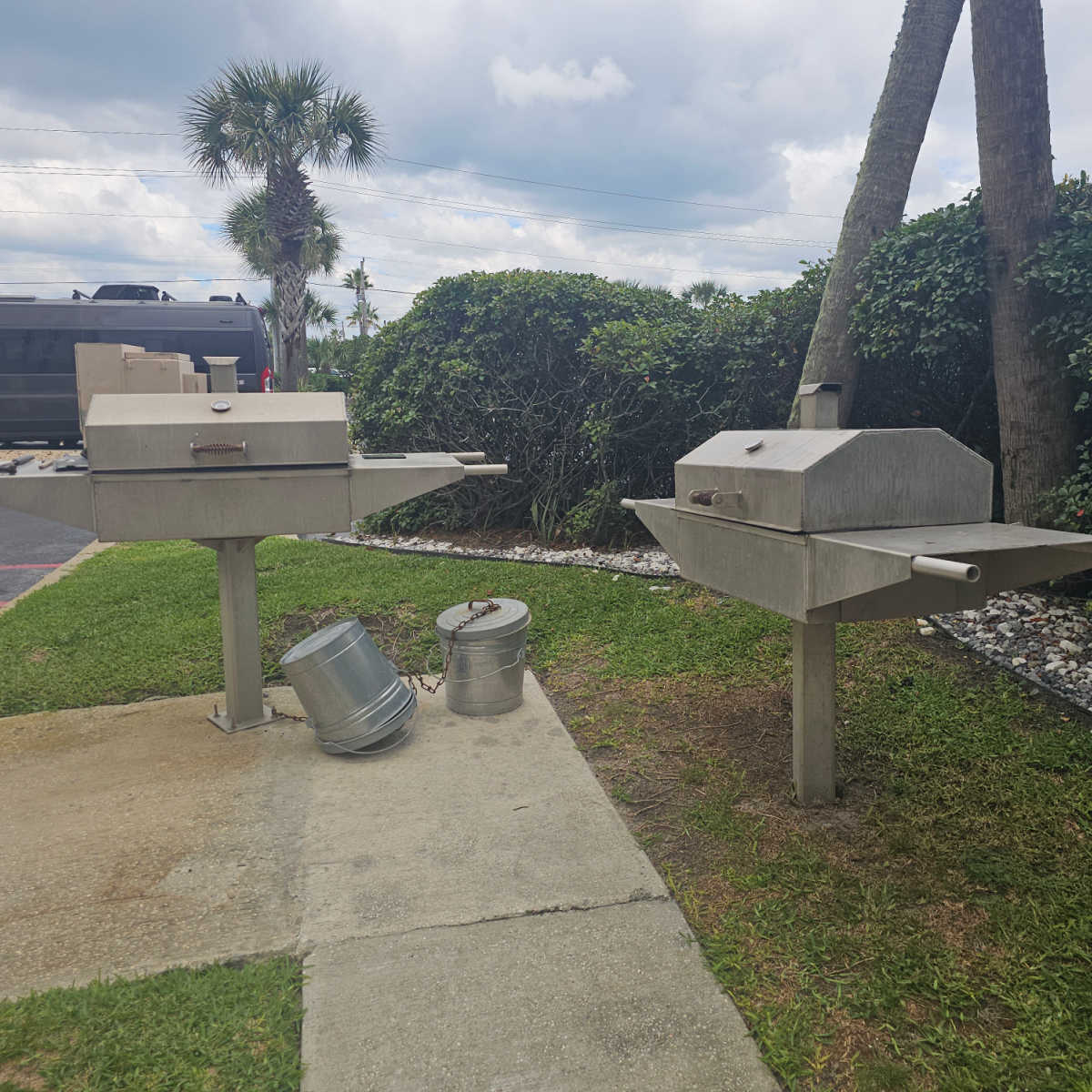 barbecue area with two barbecues, silver garbage cans under them, palm trees in the background