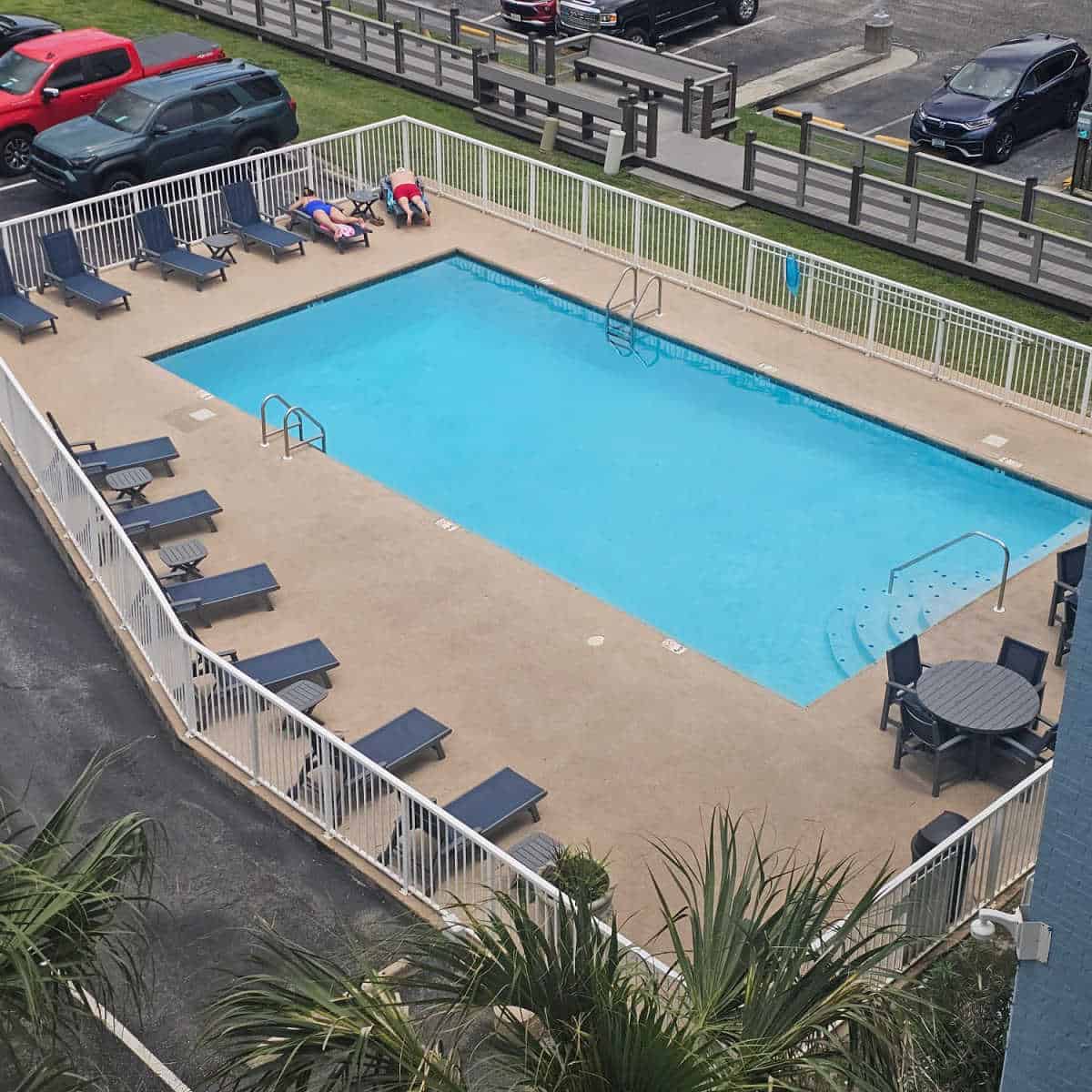 outdoor pool with lounge chairs around the pool, fenced in with cars in the background