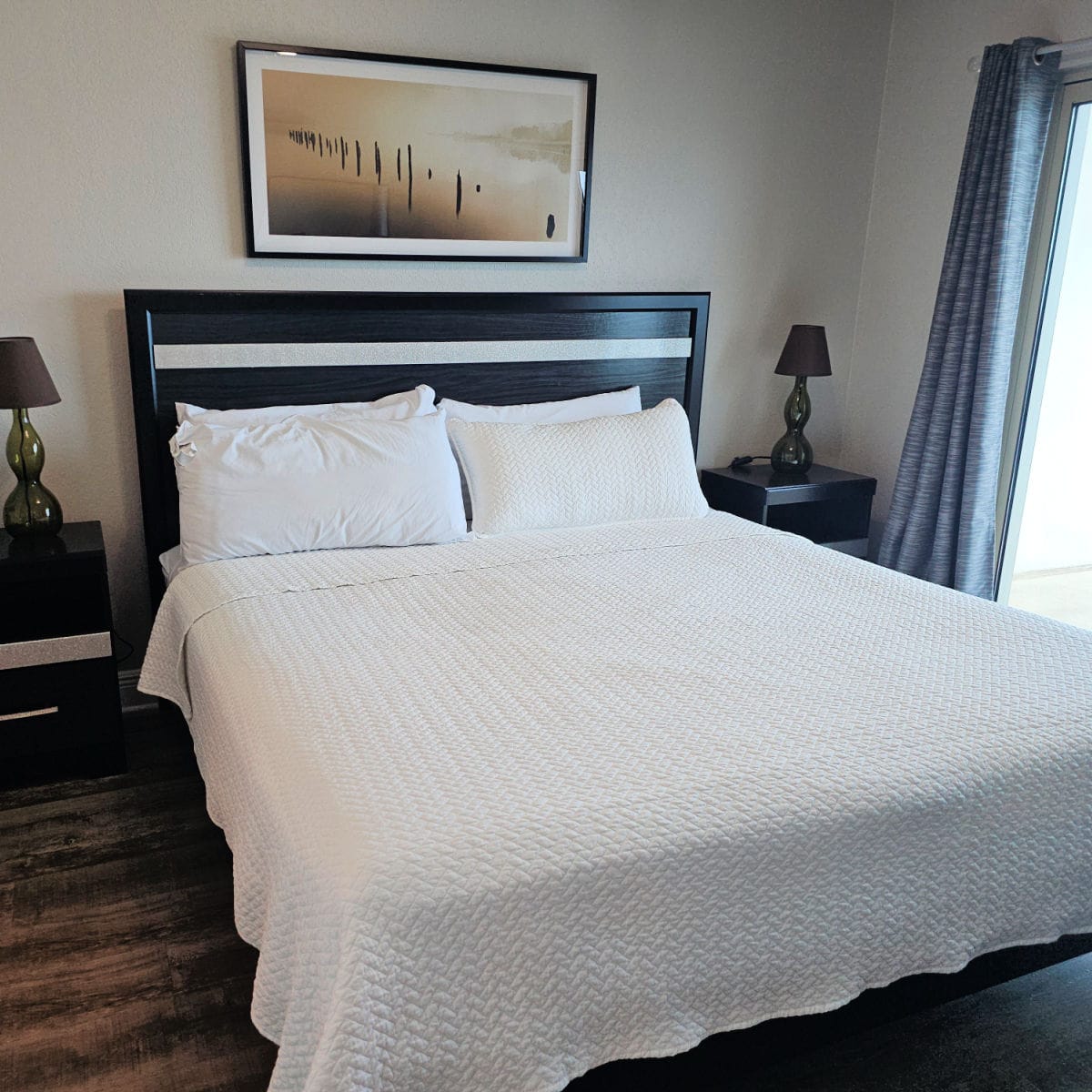bed with white linens and a dark headboard, art over the bed, lamps on a nightstand