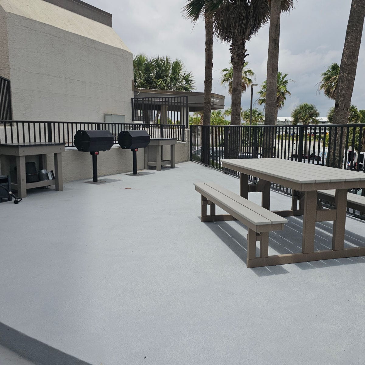 barbecue area with a picnic table, two charoal grills with small tables next to each grill, fence along the edge of the deck and palm trees behind it