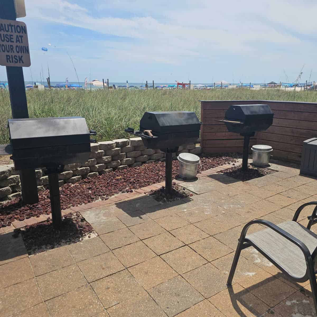 bbq area at Seawind with three barbecues in a row, views over grass to beach umbrellas and the Gulf. Corner of a chair in the lower section of the photo