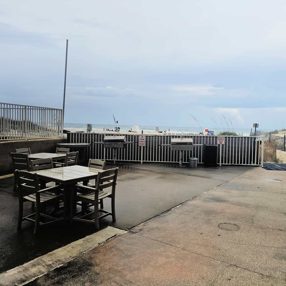 BBQ area with charcoal grills near a fence, gulf waters in the background. Table and chairs near the bbq's