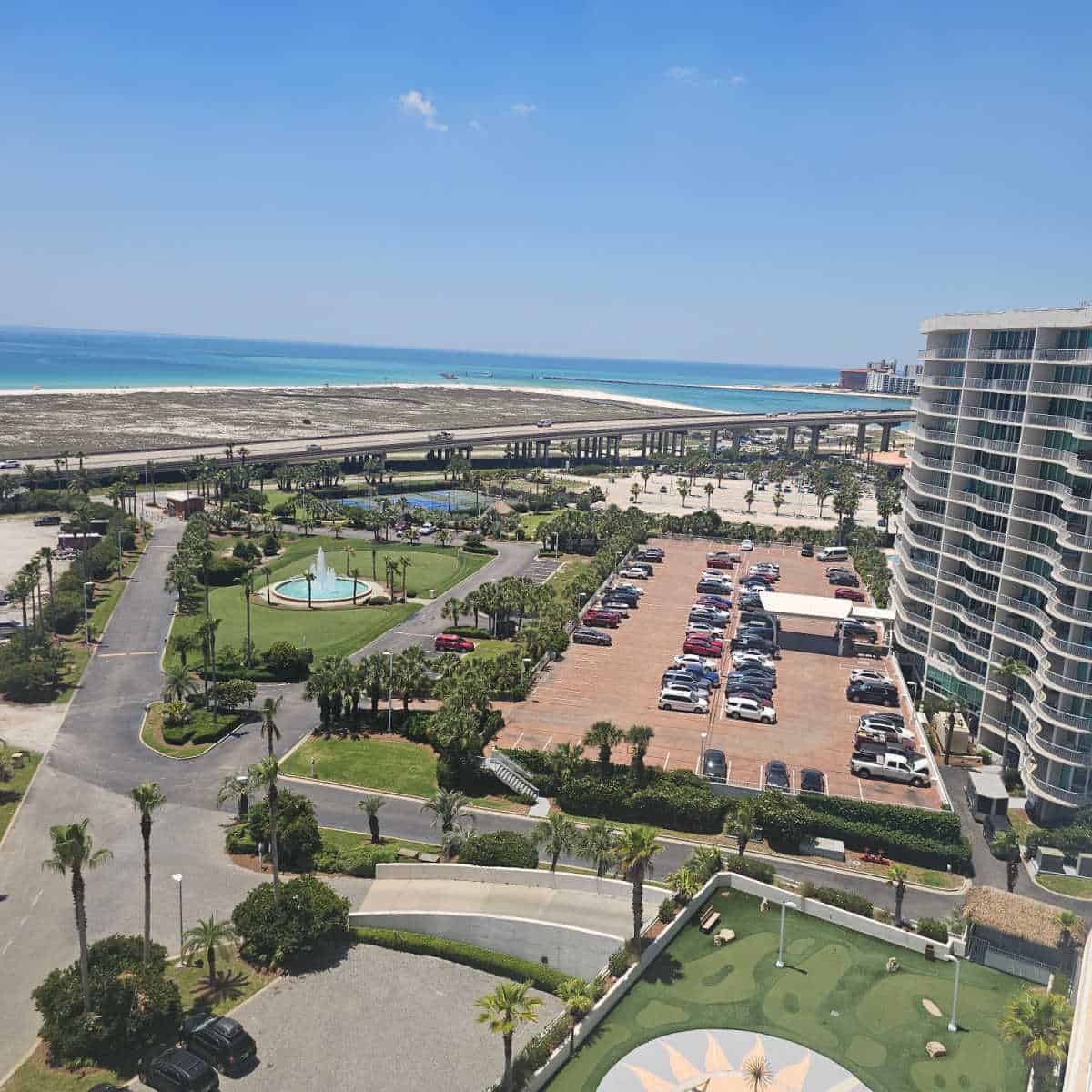 looking over the grounds of Caribe resort with parking area, putting green, Perdido Pass bridge and gulf waters in the background.