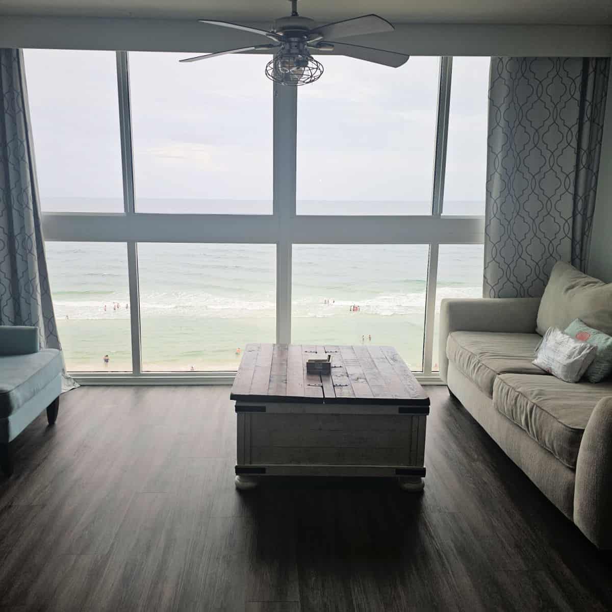 large glass window looking out to the beach, fan on the ceiling with couch and chair on the sides and a wooden table