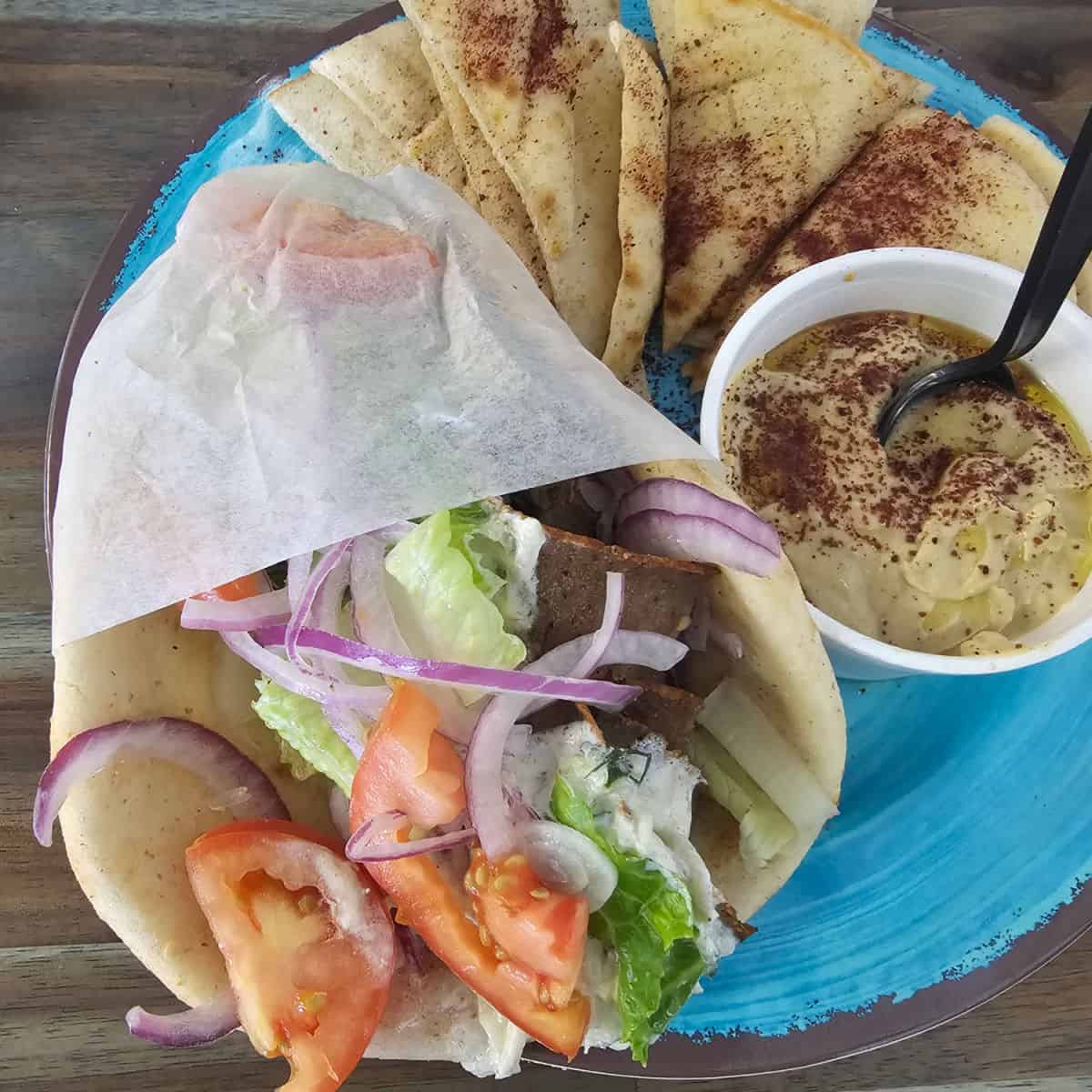 Gyro wrapped with paper on a turquoise plate with hummus in a bowl and pita triangles