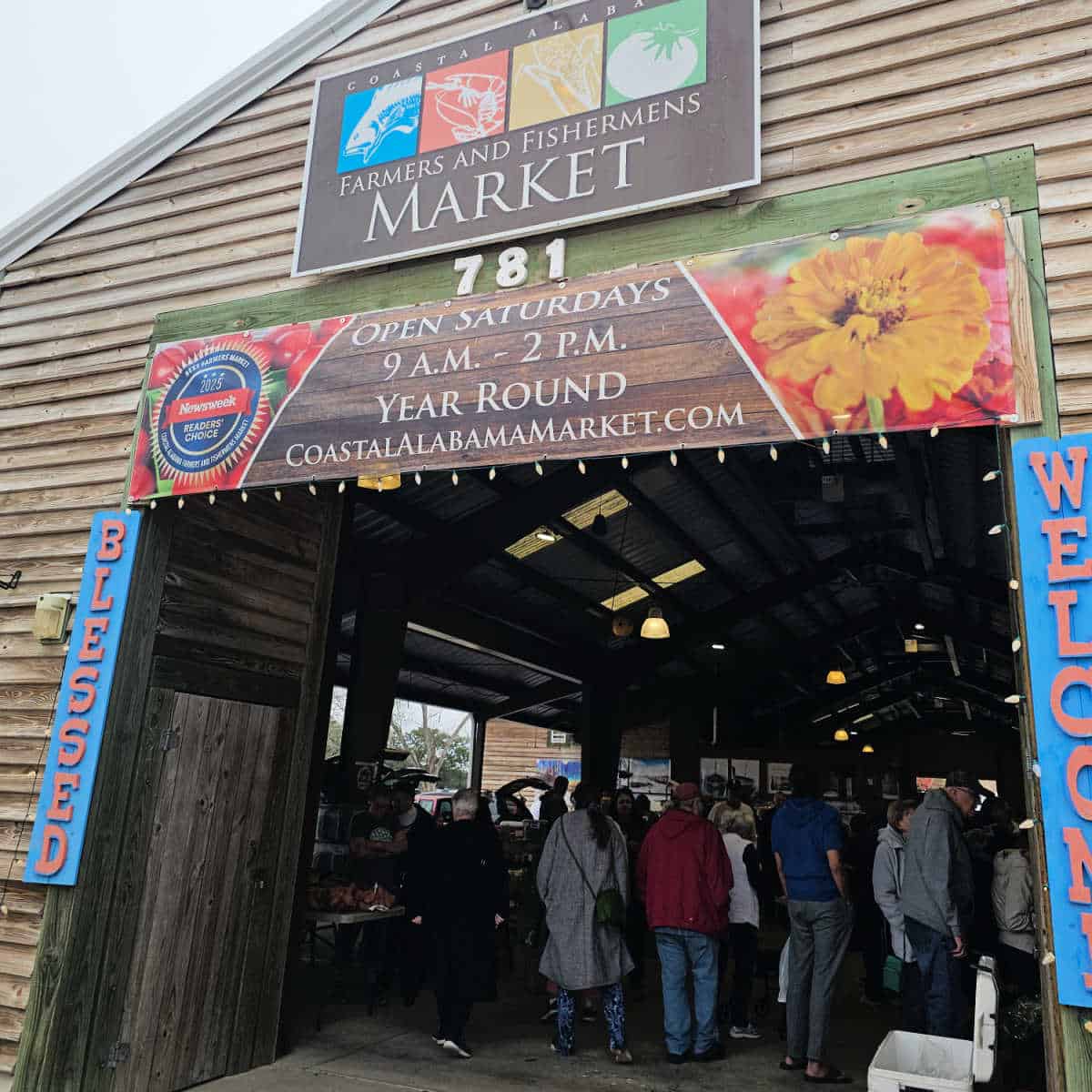 Coastal Alabama Farmers and Fishermens market sign over an open saturdays year round sign and people walking into the open air market