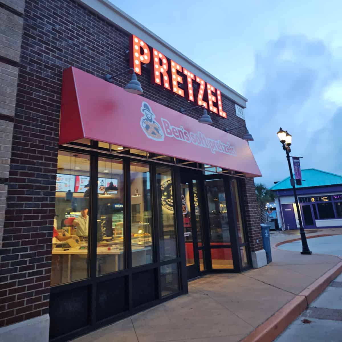 Pretzel light sign over a red awning with Ben's Soft Pretzels, windows with people working behind the windows