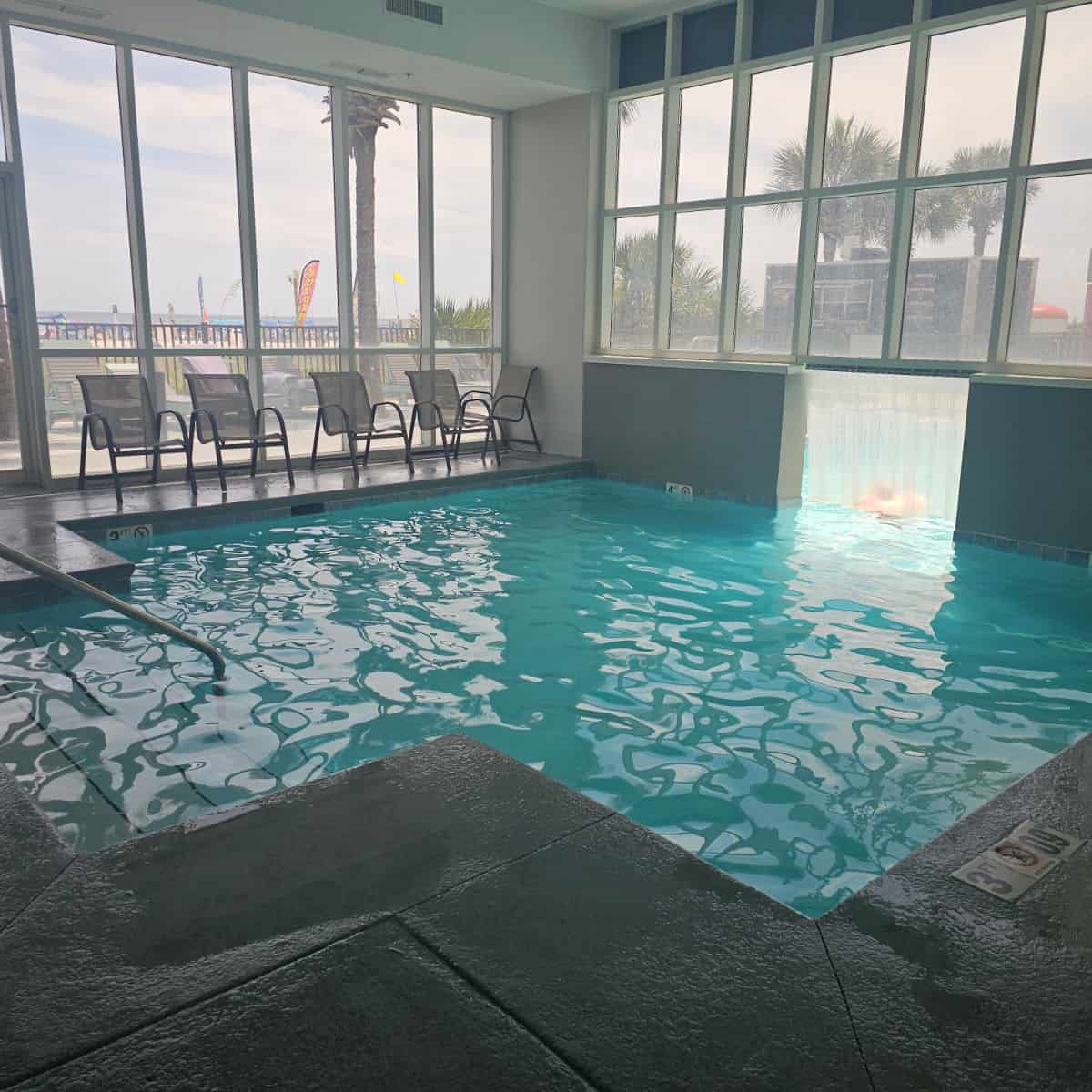 square indoor pool with chairs along the side, glass windows around the pool looking out to palm trees and the beach
