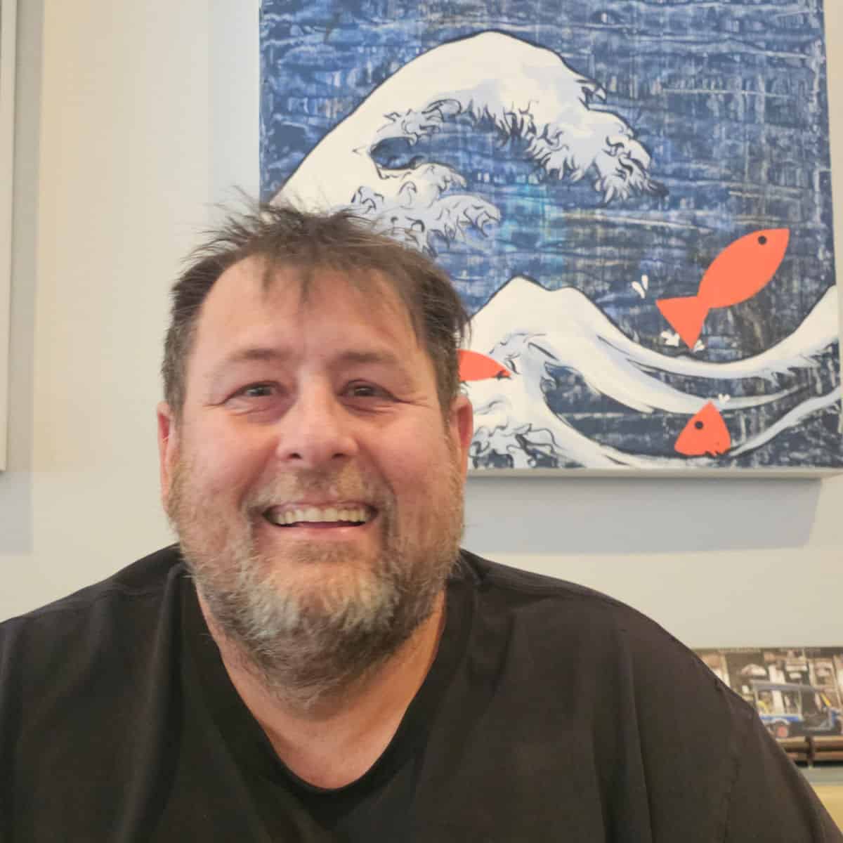 John smiling with an art print with orange fish and a wave behind him at Niki's Thai