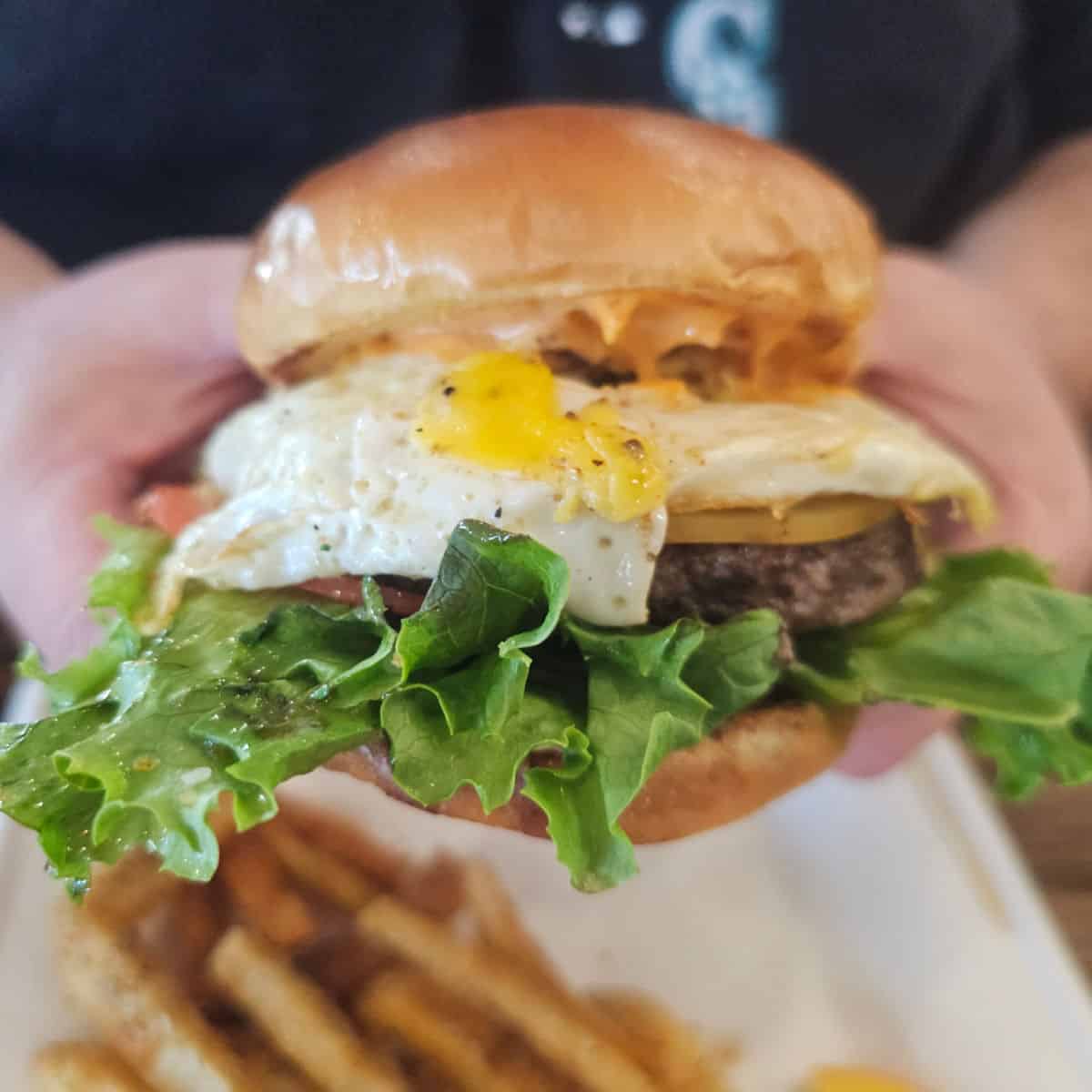 breakfast burger with runny yolk, lettuce and cheese being held above a white plate with fries