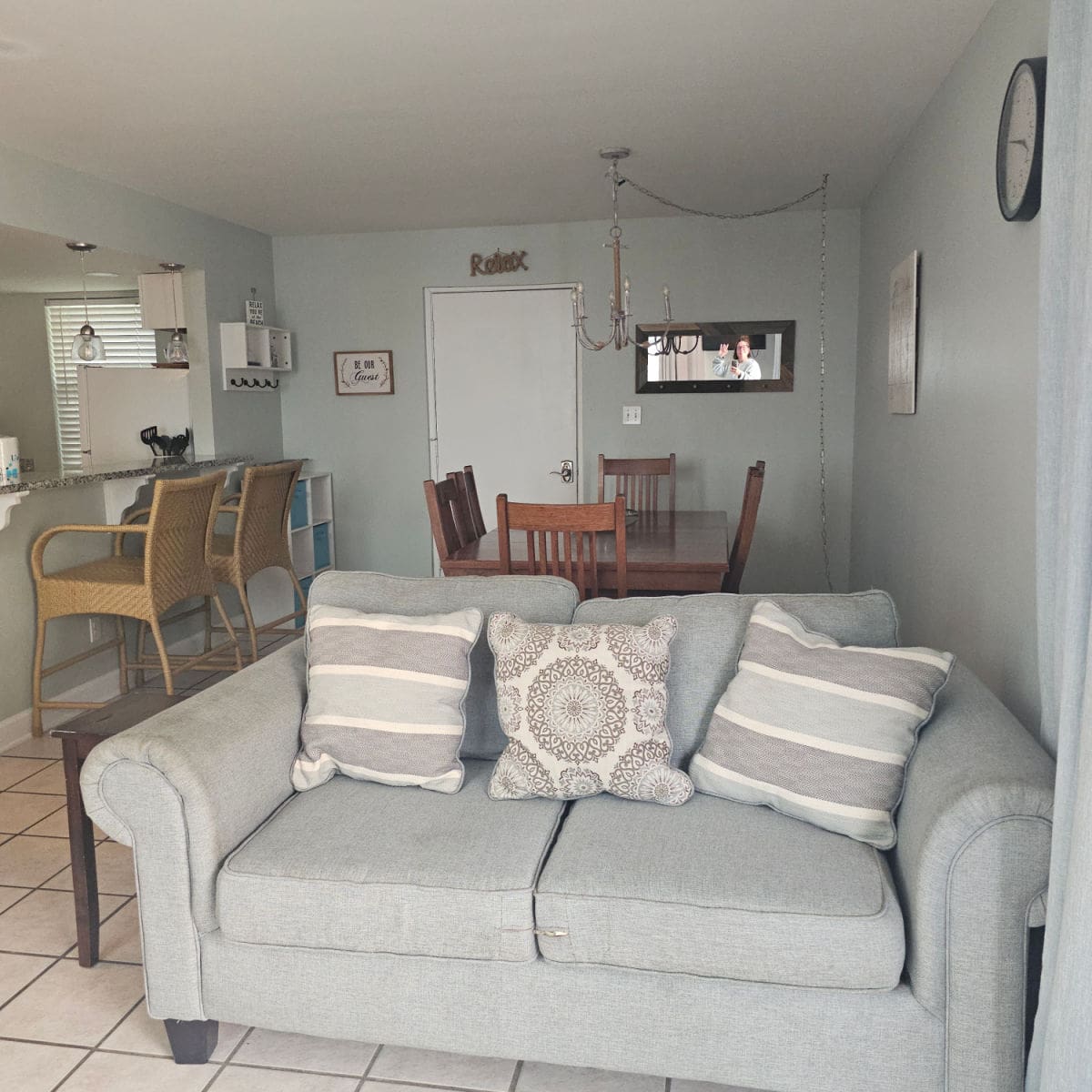Grey couch with pillows, with dining room table behind it, tammilee smiling in the mirror, kitchen on the left side with tall chairs