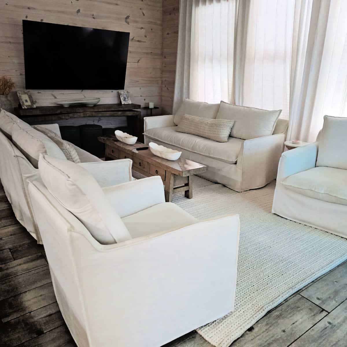 Living room with comfy white couches and chairs, brown long table between the couches, flatscreen tv on the wood paneled wall, sheer drapes over the windows