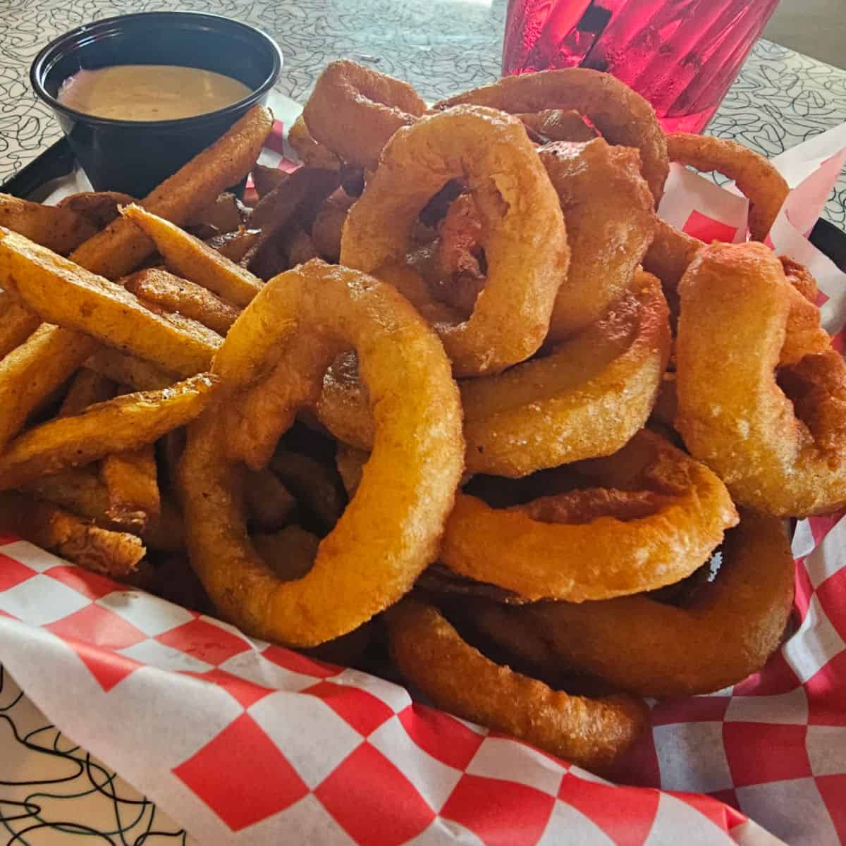 onion rings and fries in a red and white checkered paper lined basket with a cup of sauce