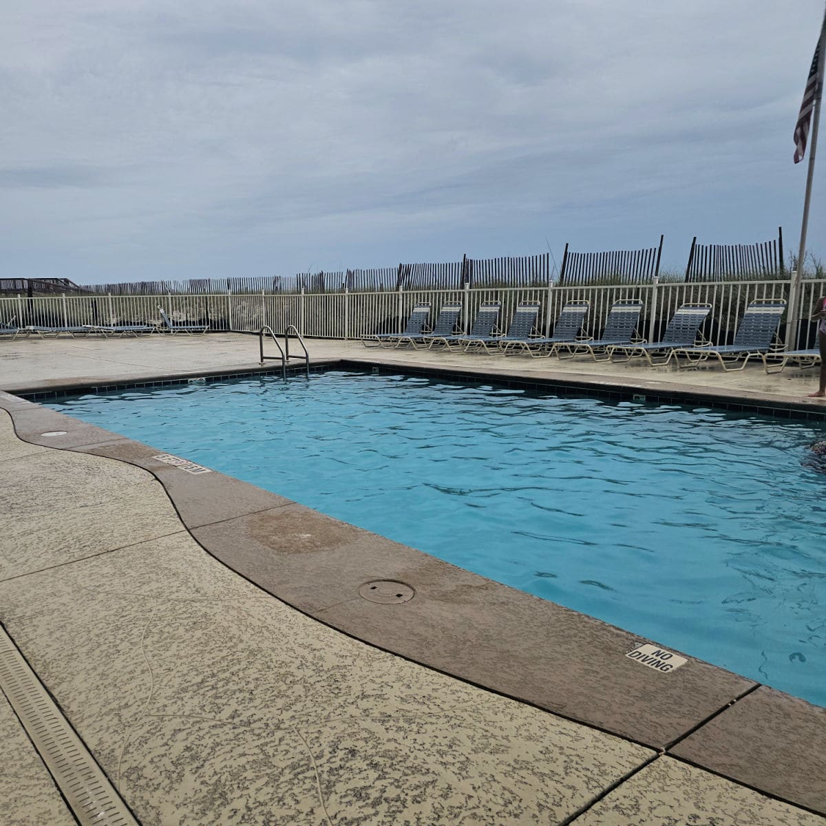 outdoor pool with lounge chairs around the edge and a fence behind the chairs leading up to a dune fence and American flag