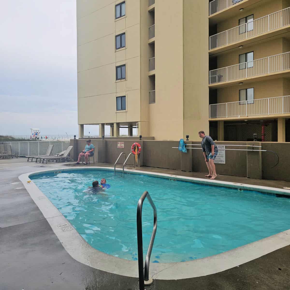 Outdoor pool with people in the water, chairs along the pool deck, tan building behind the pool