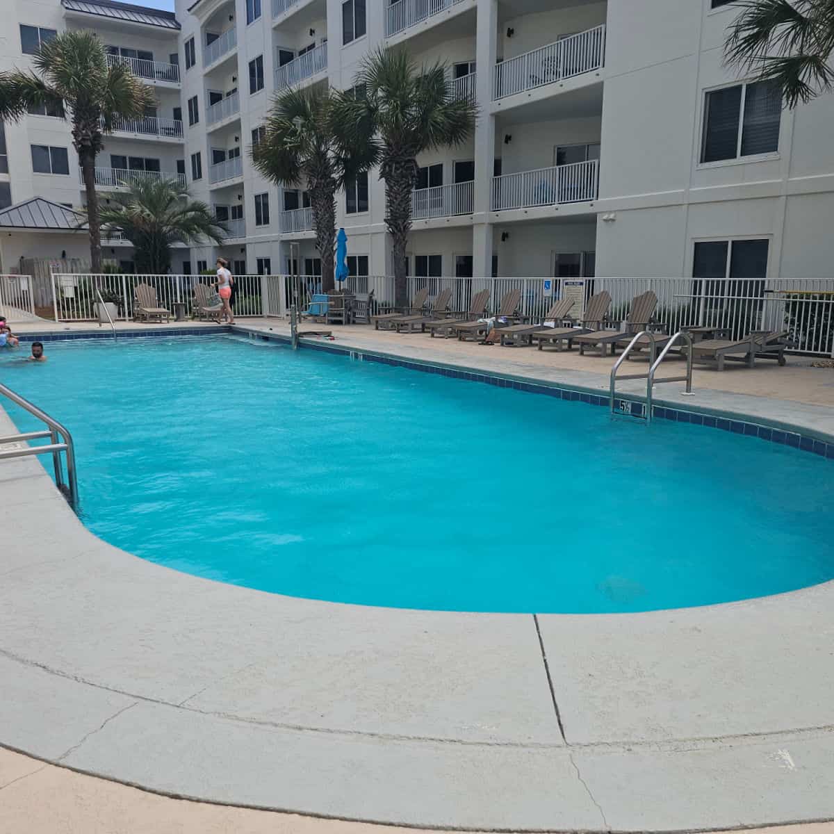 outdoor pool with deck chairs around it, fence surrounding the pool deck, white buildings behind the fence