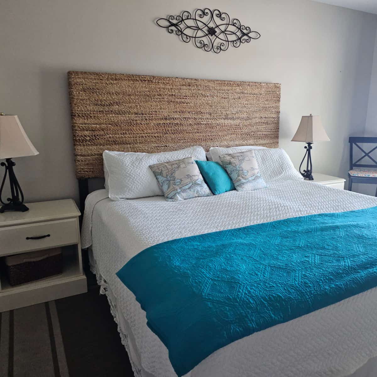 bedroom with king size bed with white linens and turquoise throw and world pillows with a rattan headboard, metal art above the bed, nightstands with lamps, and a chair in the corner