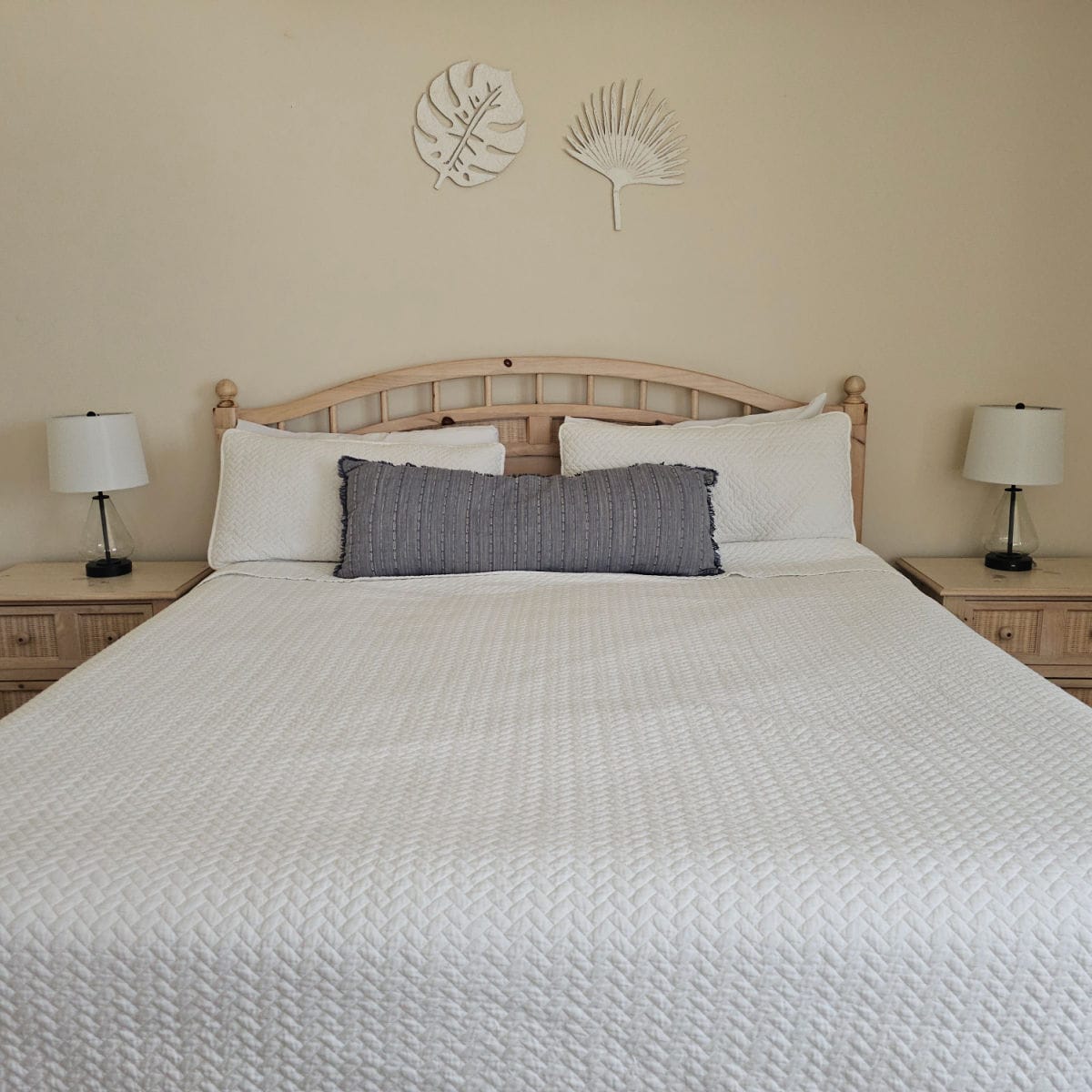King size bed with a white blanket, dark pillow in front of white pillows, two nightstands with matching glass and white shaded lamps, and two leaf art pieces on the wall above the bed