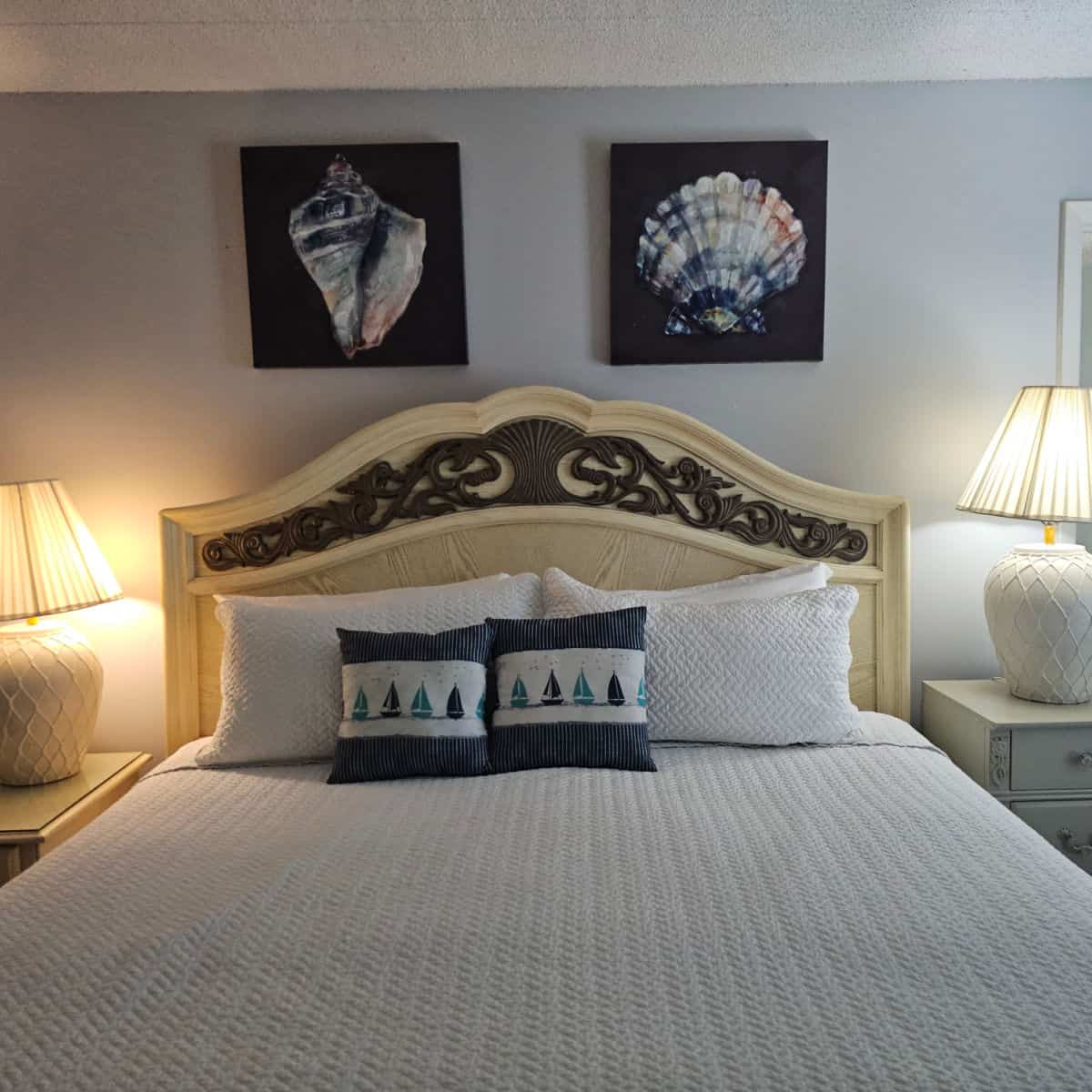 Bedroom with white linens on the bed and a couple of pillows with sailboats on them, shell images over the bed. Nightstands with large lamps on either side of the bed.