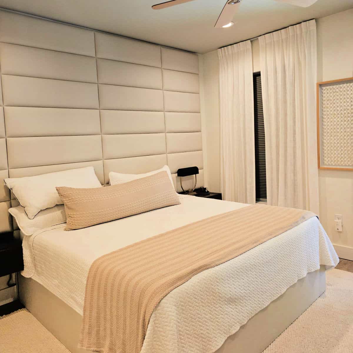 Bedroom with a padded cream colored wall, bed with tan and white linens, art on the wall