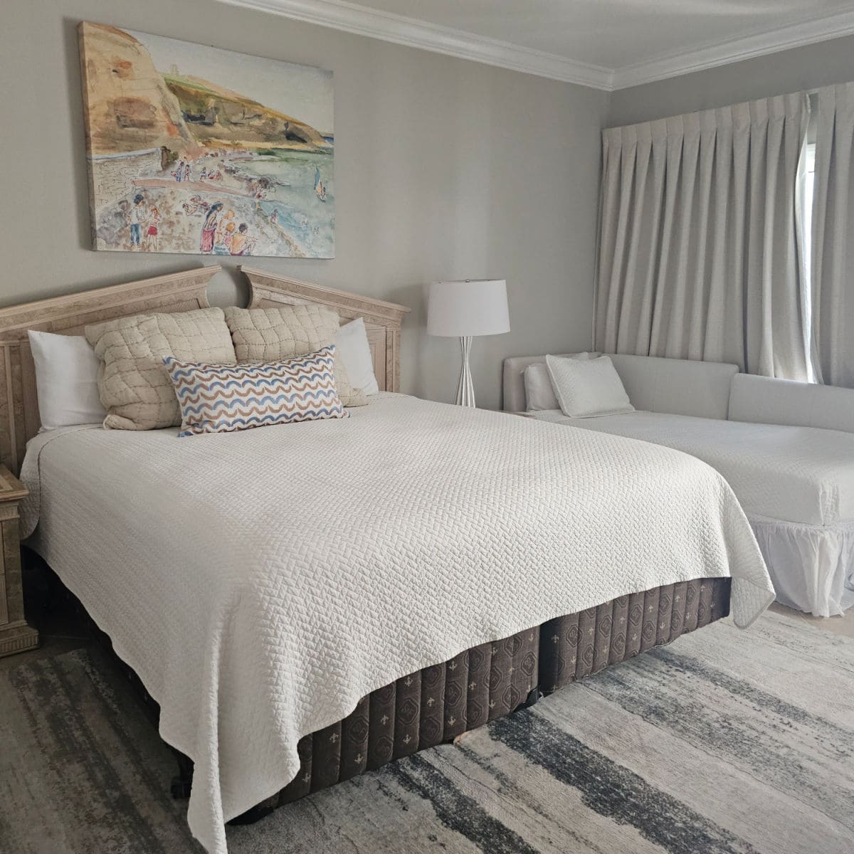 bedroom with a king bed covered in a coverlet and multiple pillows, art print above the bed, small couch bed to the side of the bed with white curtains over the window