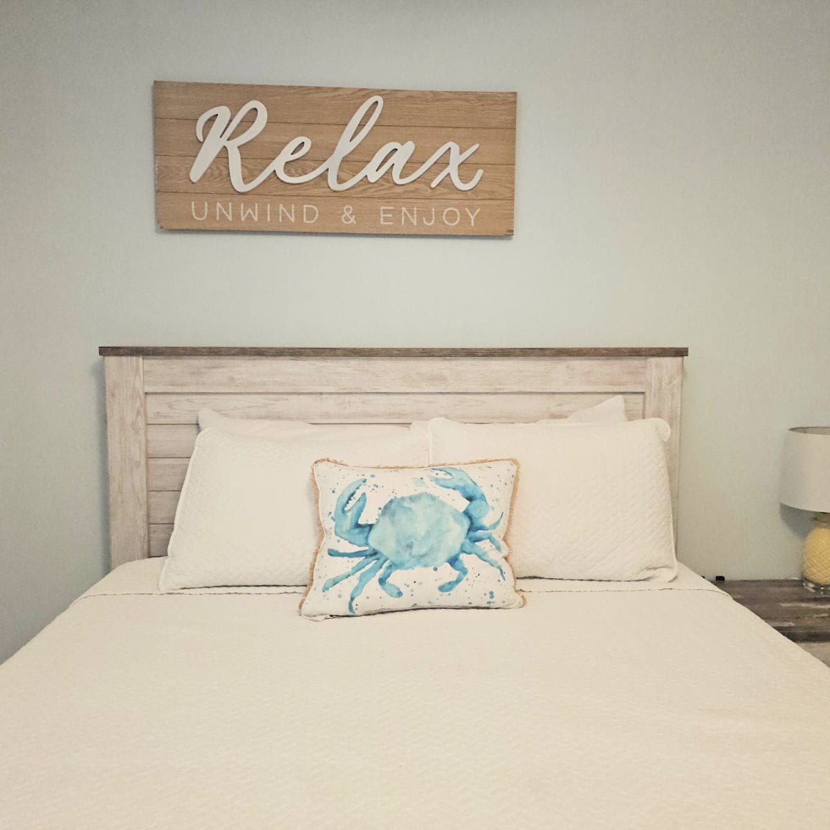 Relax Unwind & Enjoy wooden sign over a bed with white pillows and a crab pillow, lamp to the side of the bed
