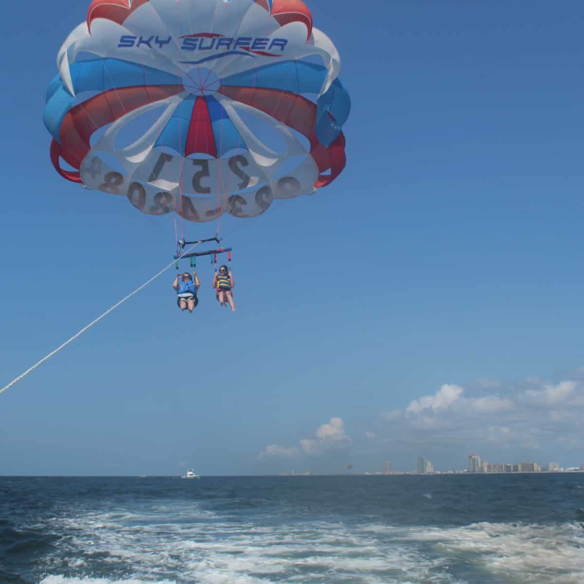 Skysurfer red white and blue parasail with tammilee and amy below it floating over the water with Orange Beach in the background