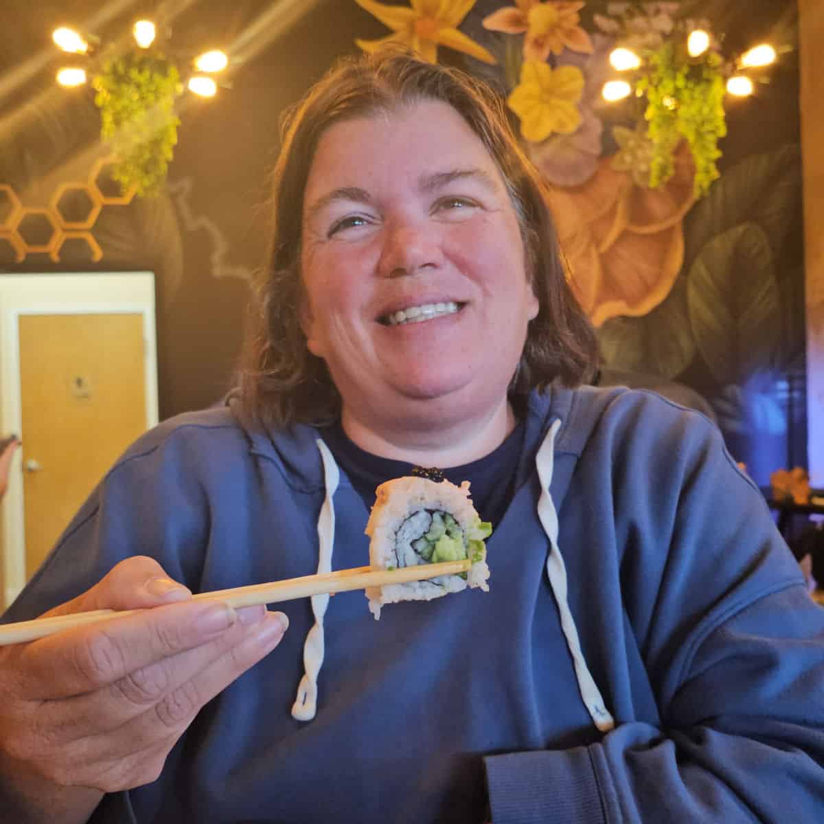 tammilee holding a piece of vegan sushi in chopsticks with a colorful wall behind her