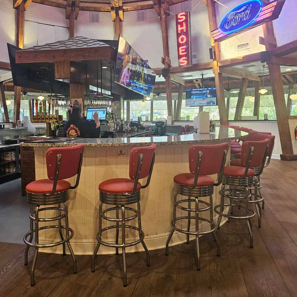bar stools around a curved bar inside the restaurant with flatscreen tv's over the bar, a ford neon sign and SHOES neon sign, women at a computer touching the screen in the middle of the bar