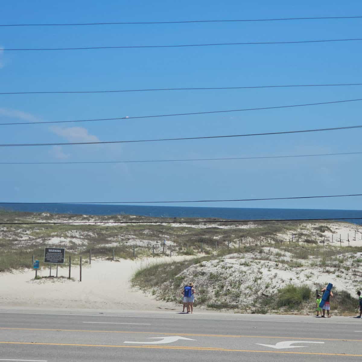 looking across the road to people with beach gear walking to a sand path leading to the beach