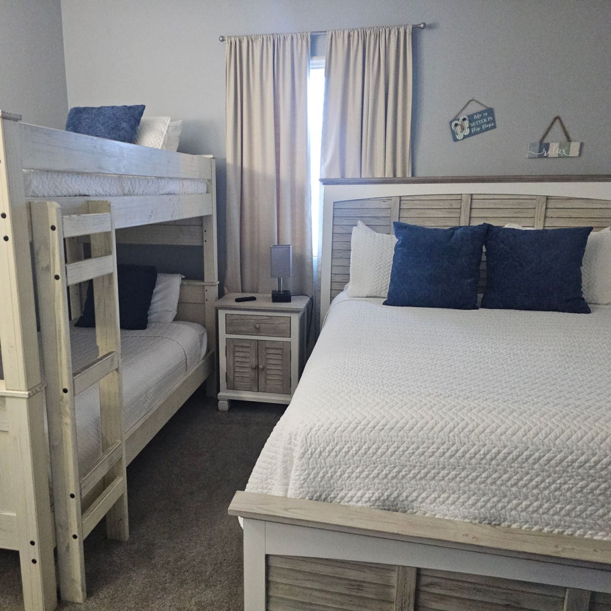 bedroom with a bunkbed on the left side, night stand and then bed with white linens, two blue square pillows, small art pieces on the wall above the bed