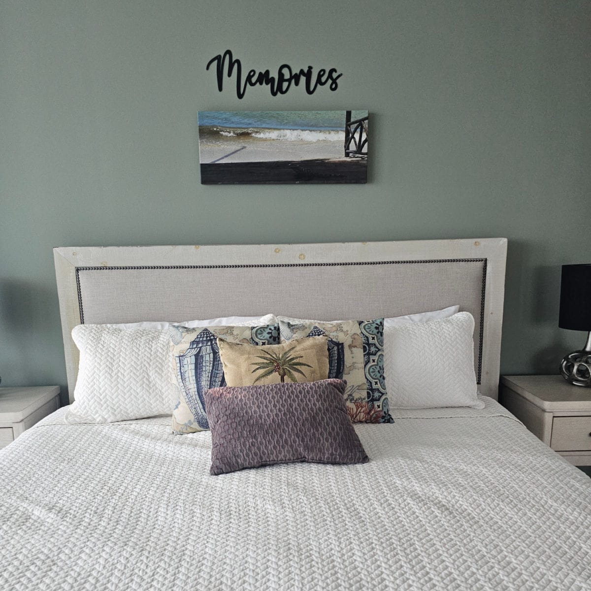 Bedroom with memories sign and a art photo on the wall, bed with padded headboard, white linens, and colorful pillows, nightstand next to the bed with lamp and dark lampshade