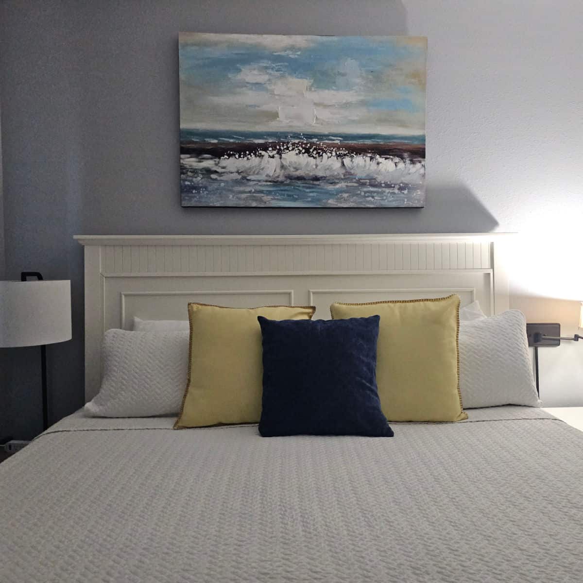 bedroom with bed with white linens, yellow and blue pillows, nightstand with lamp, art over the bed with a coastal scene