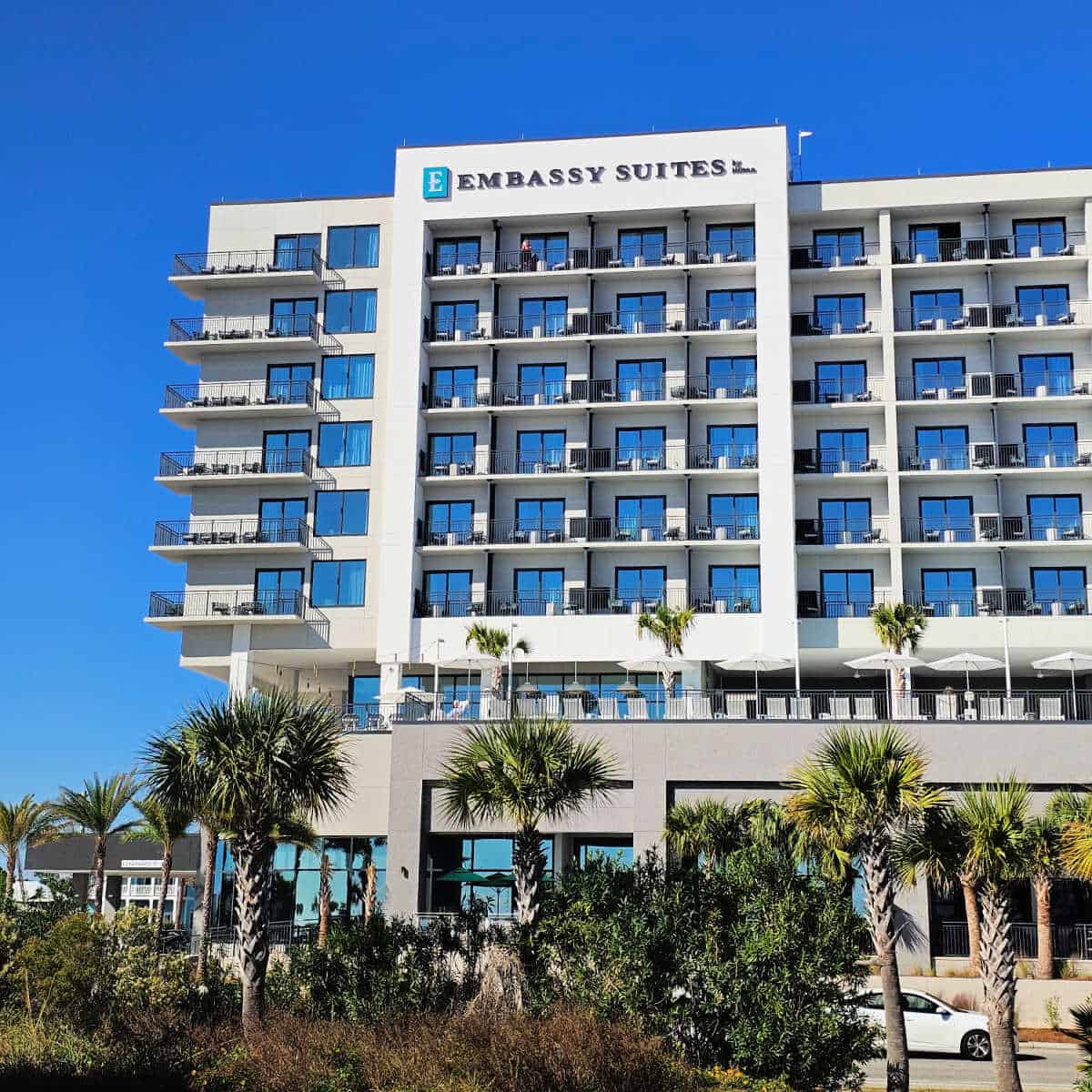 Embassy Suites by Hilton sign o the top of the hotel with 6 floors of rooms with balconys, a pool deck, and palm trees along the road.