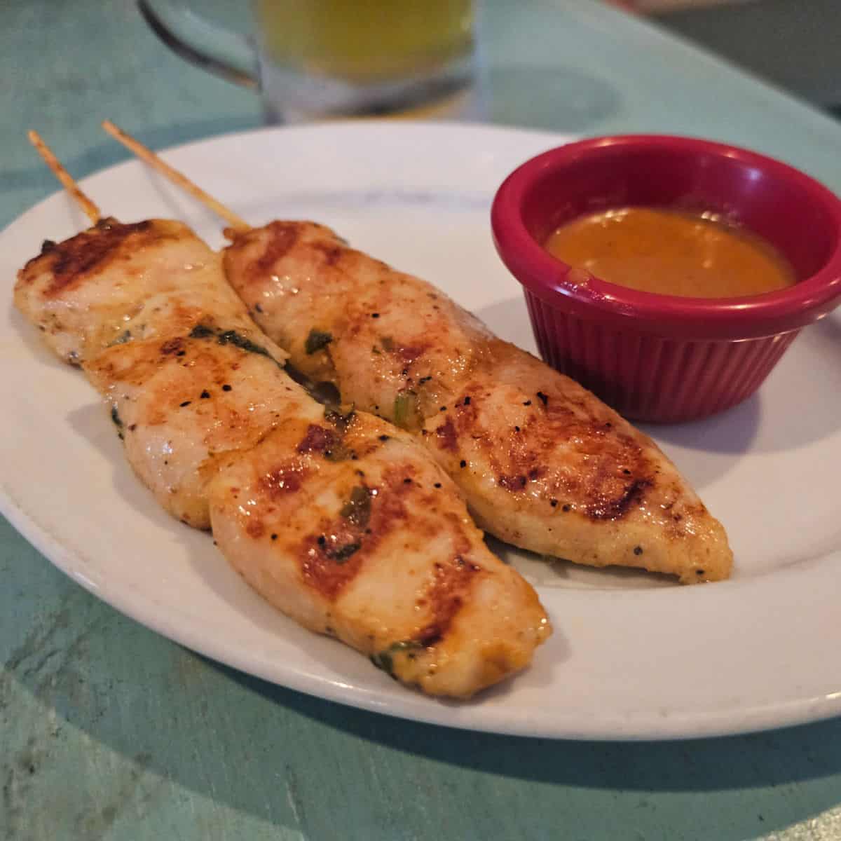 chicken satay on skewers on a white plate with a jar of sauce