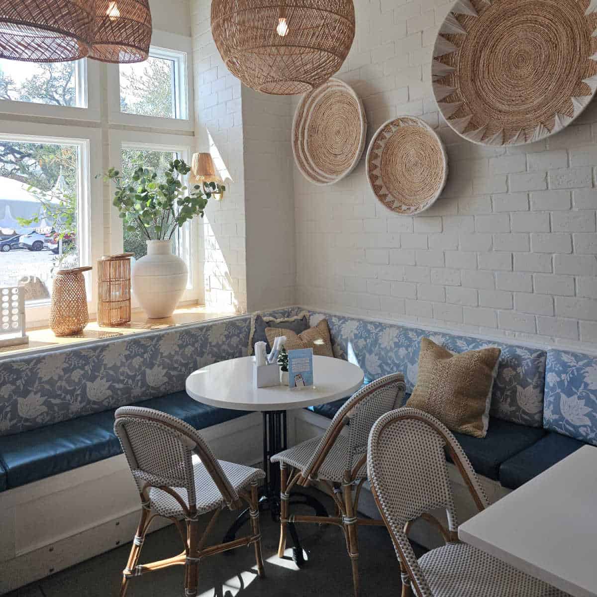 corner seating with a white and blue wrap around bench with padded seat, white table, chairs, baskets on the brick wall, vases and lanterns in the window