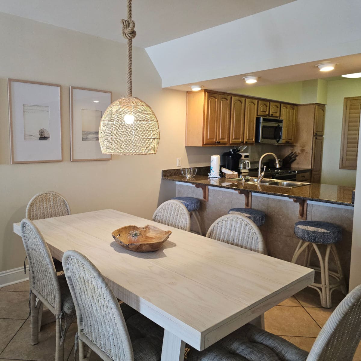 Dining room area with a wood table surrounded by wicker chairs, wicker lamp hanging over the table, three bar stools under the kitchen counter as you look towards the kitchen area with wood cabinets and stainless steel appliances