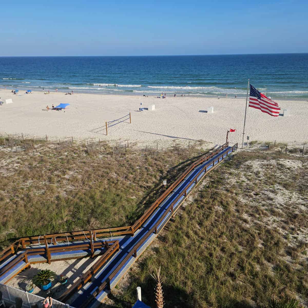 wooden boardwalk over sand dunes to the beach with a large American flag, sandy area, then Gulf waters