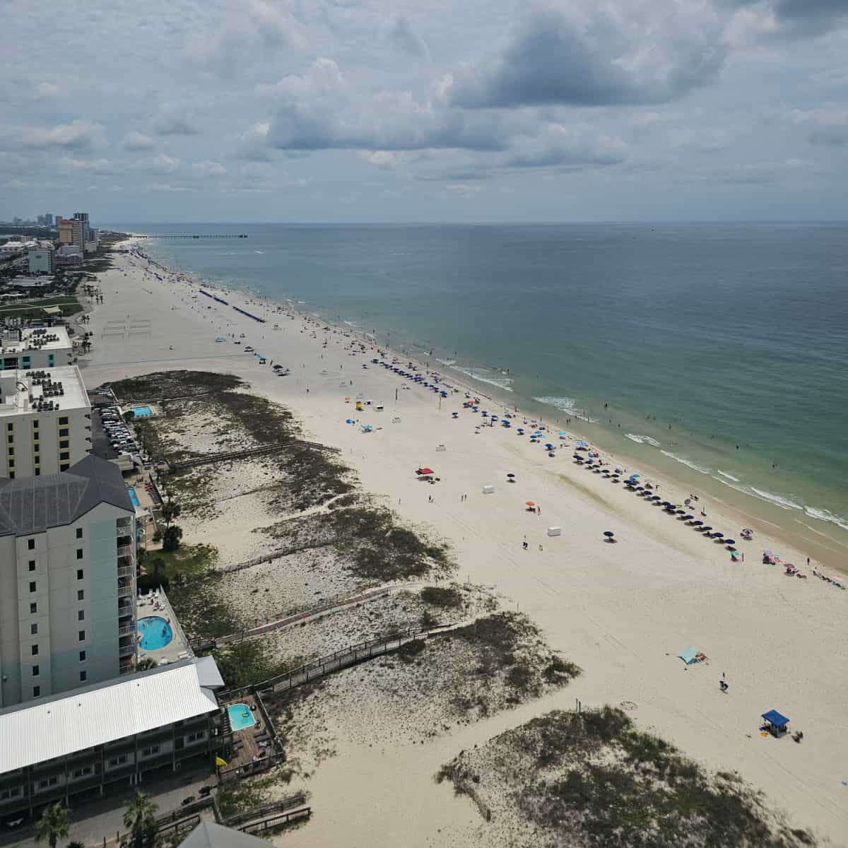 looking down over the beach with Gulf Shores condos in a row leading to the pier, sand dunes and beach umbrellas along the waters shoreline