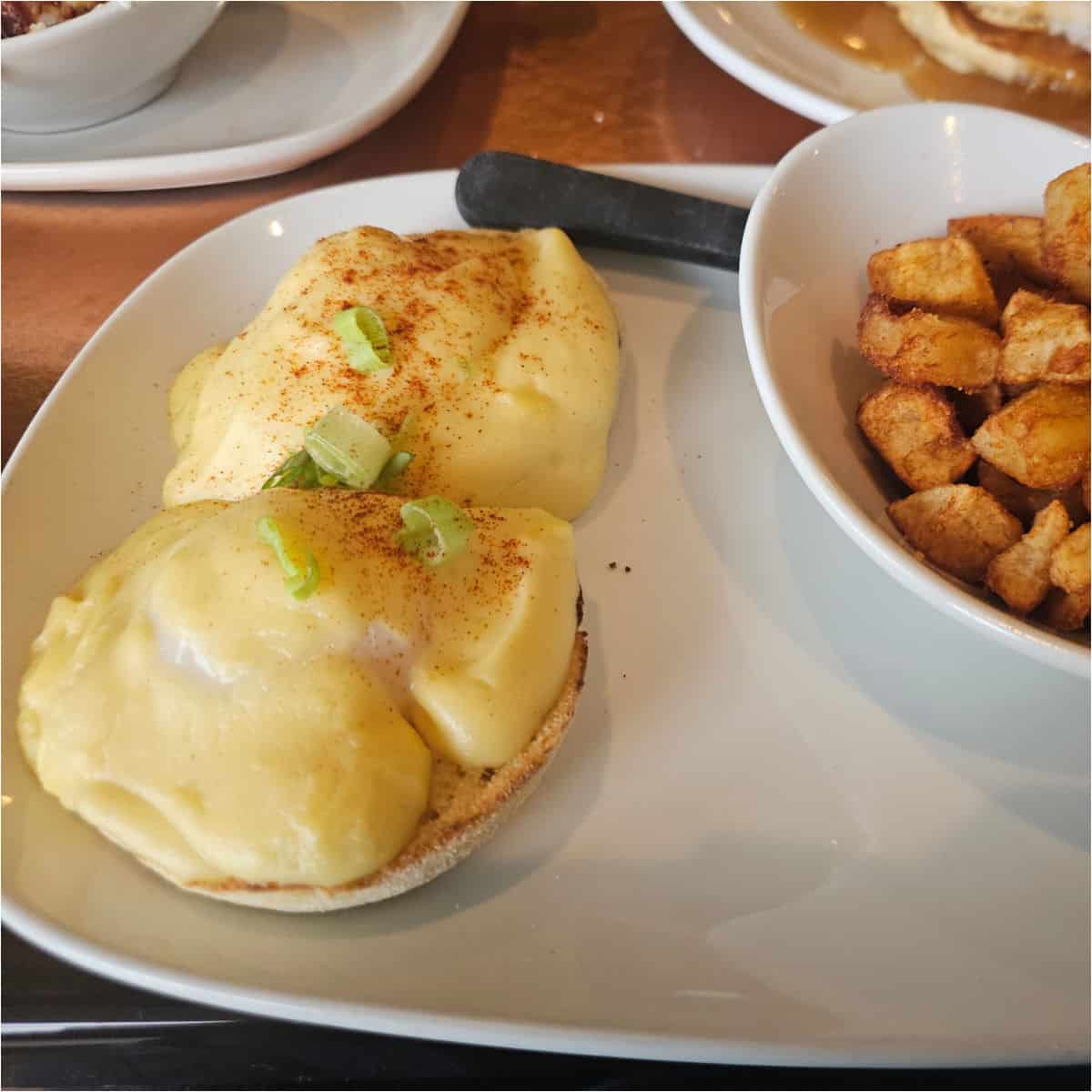Eggs benedict on a white plate with a bowl of home fries